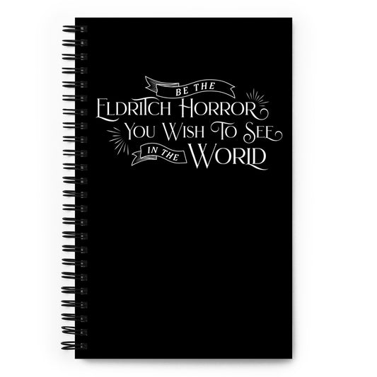 A black wire-bound notebook featuring white, semi-gothic style text that reads "Be the Eldritch Horror you wish to see in the world".