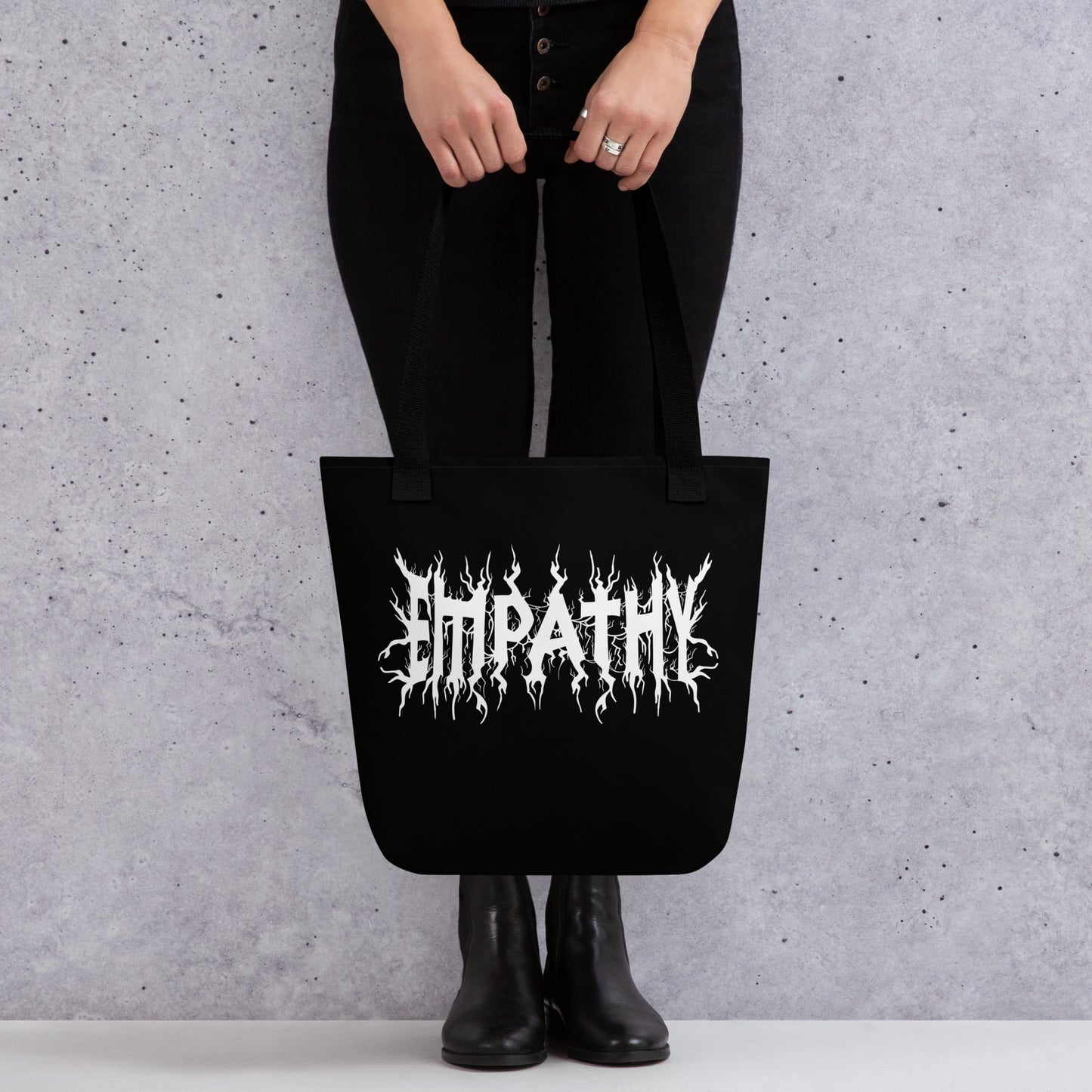A waist-down image of a model wearing all black and holding a tote bag. The tote bag is black and features white text reading "Empathy" in the jagged style of heavy metal band shirts