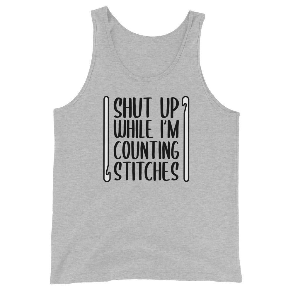 A grey tank top featuring black text that reads "Shut up while I'm counting stitches." The text is framed by a crochet hook to the left and right.