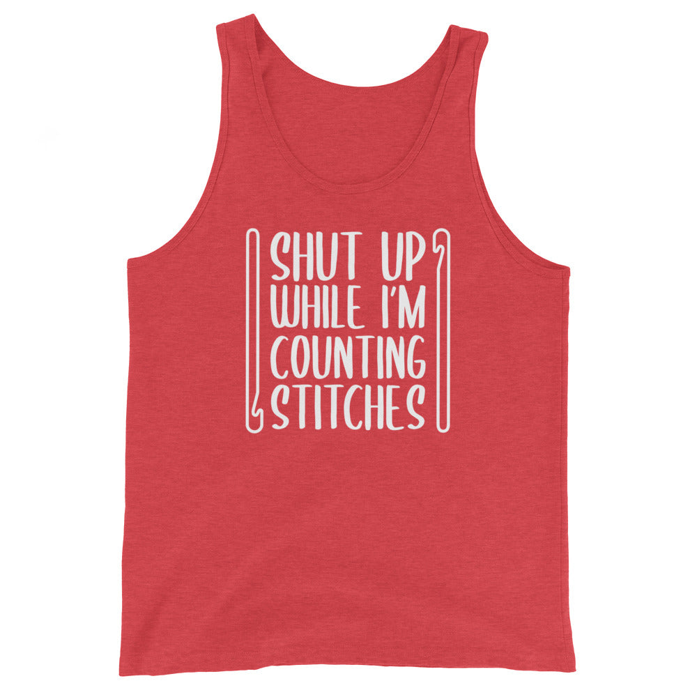 A red tank top featuring white text that reads "Shut up while I'm counting stitches." The text is framed by a crochet hook to the left and right.