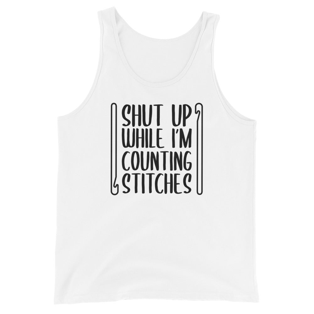 A white tank top featuring black text that reads "Shut up while I'm counting stitches." The text is framed by a crochet hook to the left and right.