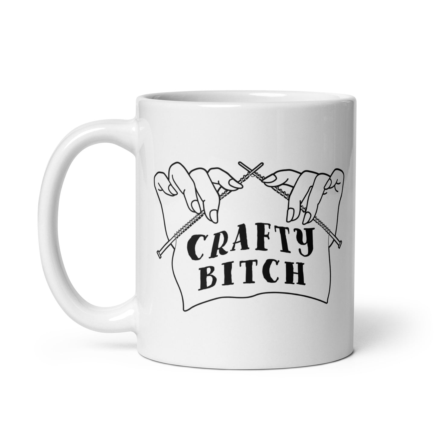 A white ceramic 11 ounce coffee mug featuring a single-color illustration of a pair of hands holding knitting needles. Fabric on the needles features text that reads "crafty bitch".