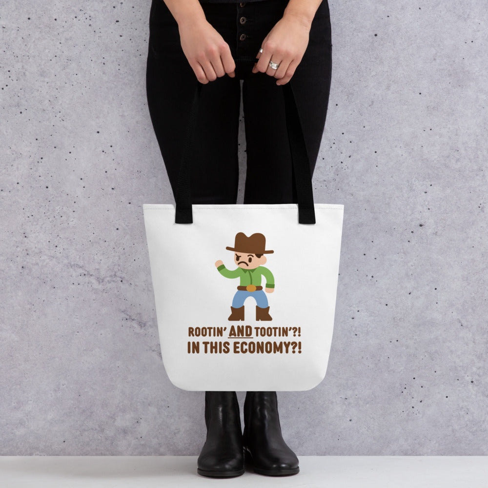 A waist-down image of a model wearing all black and holding a tote bag. The tote bag is a white canvas bag with black handles and features an illustration of a confused-looking cowboy wearing a green shirt. Text underneath the cowboy reads "Rootin' AND tootin'?! In this economy?!"