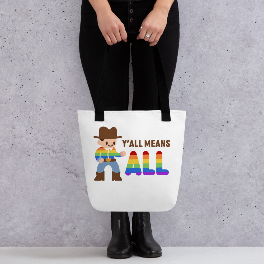 A waist-down image of a model wearing all black. The model is holding a white canvas tote bag with black handles featuring an illustration of a smiling cowboy wearing a rainbow striped shirt. Text alongside the cowboy reads "Y'all means ALL". The word "ALL" is rainbow-colored to match the cowboy's shirt.