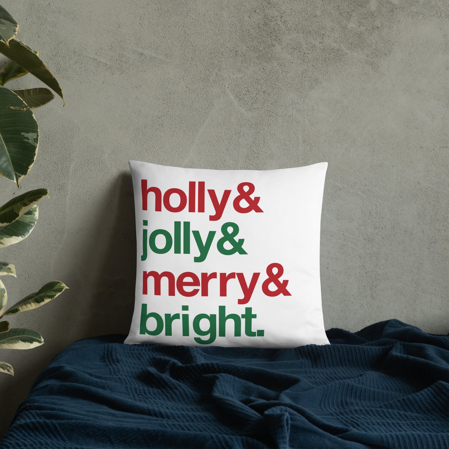 An 18" by 18" throw pillow decorated with four lines of red and green text that read "Holly & jolly & merry & bright". The pillow is set on top of a bed with a blue blanket against a concrete wall.