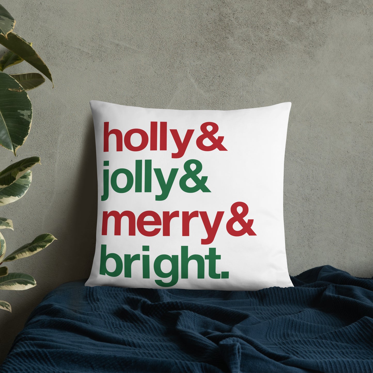 A 22" by 22" throw pillow decorated with four lines of red and green text that read "Holly & jolly & merry & bright". The pillow is set on top of a bed with a blue blanket against a concrete wall.