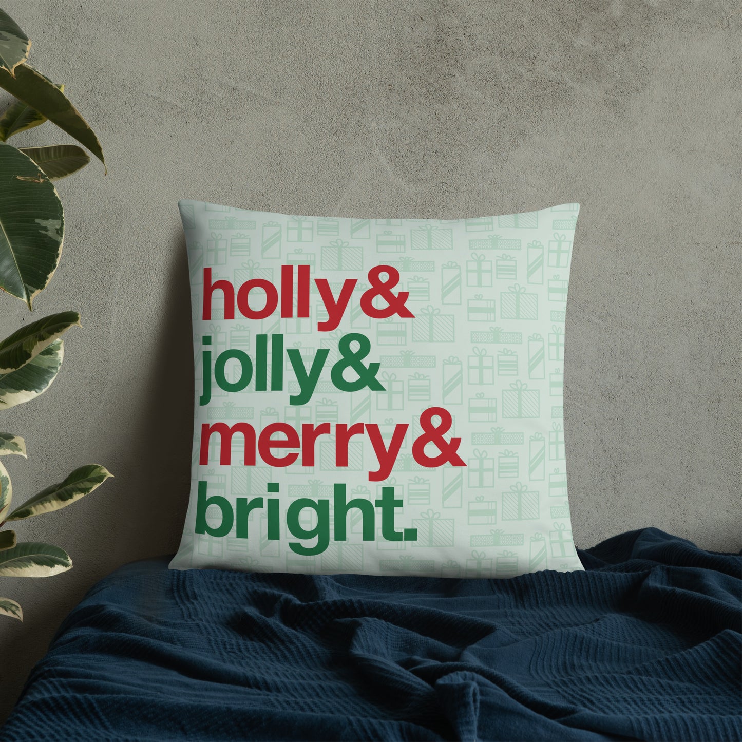 A 22" by 22" throw pillow decorated with a pale green background pattern of presents. There are also four lines of red and green text on the pillow that read "Holly & jolly & merry & bright". The pillow is set on top of a bed with a blue blanket against a concrete wall.