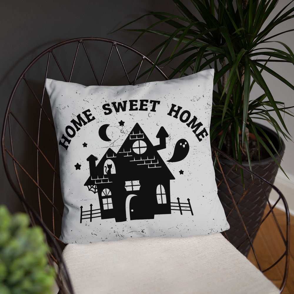A white throw pillow on a metal chair, surrounded by houseplants. The pillow has a black illustration of a haunted house on it and text reading "home sweet home".
