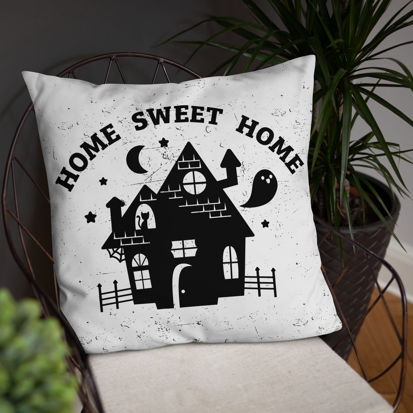 A white throw pillow on a metal chair, surrounded by houseplants. The pillow has a black illustration of a haunted house on it and text reading "home sweet home".