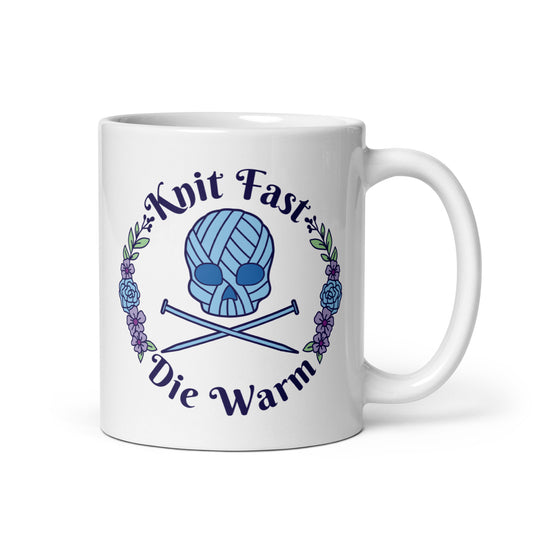 A white 11 ounce mug featuring an image of a blue skull and crossbones made of yarn and knitting needles. A floral wreath surrounds the skull, along with words that read "Knit Fast, Die Warm"