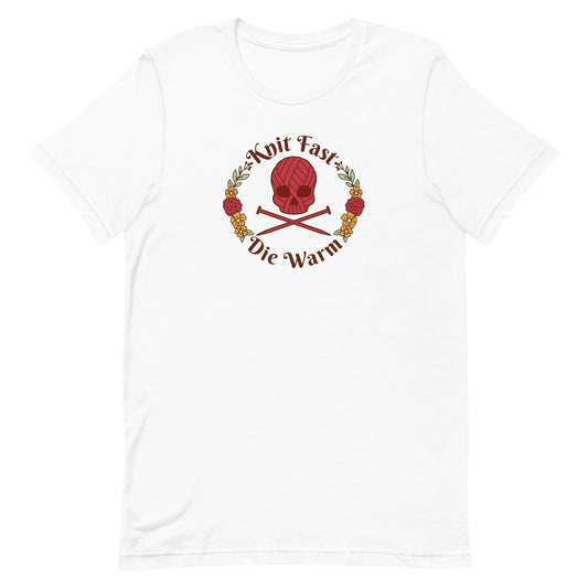 A white crewneck t-shirt featuring an image of a skull and crossbones made of yarn and knitting needles. A floral wreath surrounds the skull, along with words that read "Knit Fast, Die Warm"