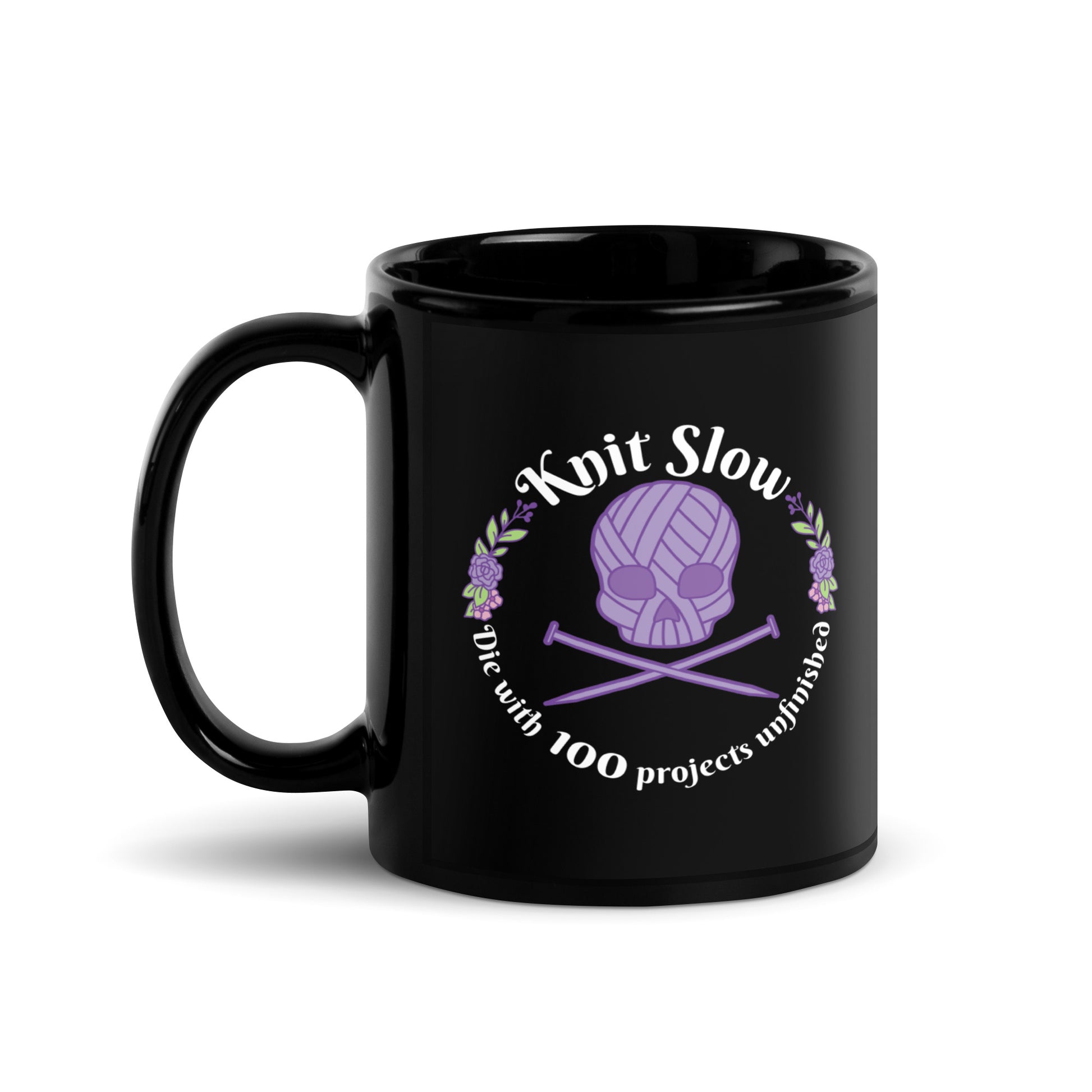 A black 11 ounce ceramic mug featuring an image of a skull and crossbones made of yarn and knitting needles. A floral wreath surrounds the skull, along with words that read "Knit Slow, Die with 100 projects unfinished"