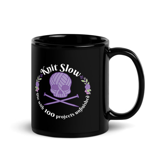 A black 11 ounce ceramic mug featuring an image of a skull and crossbones made of yarn and knitting needles. A floral wreath surrounds the skull, along with words that read "Knit Slow, Die with 100 projects unfinished"