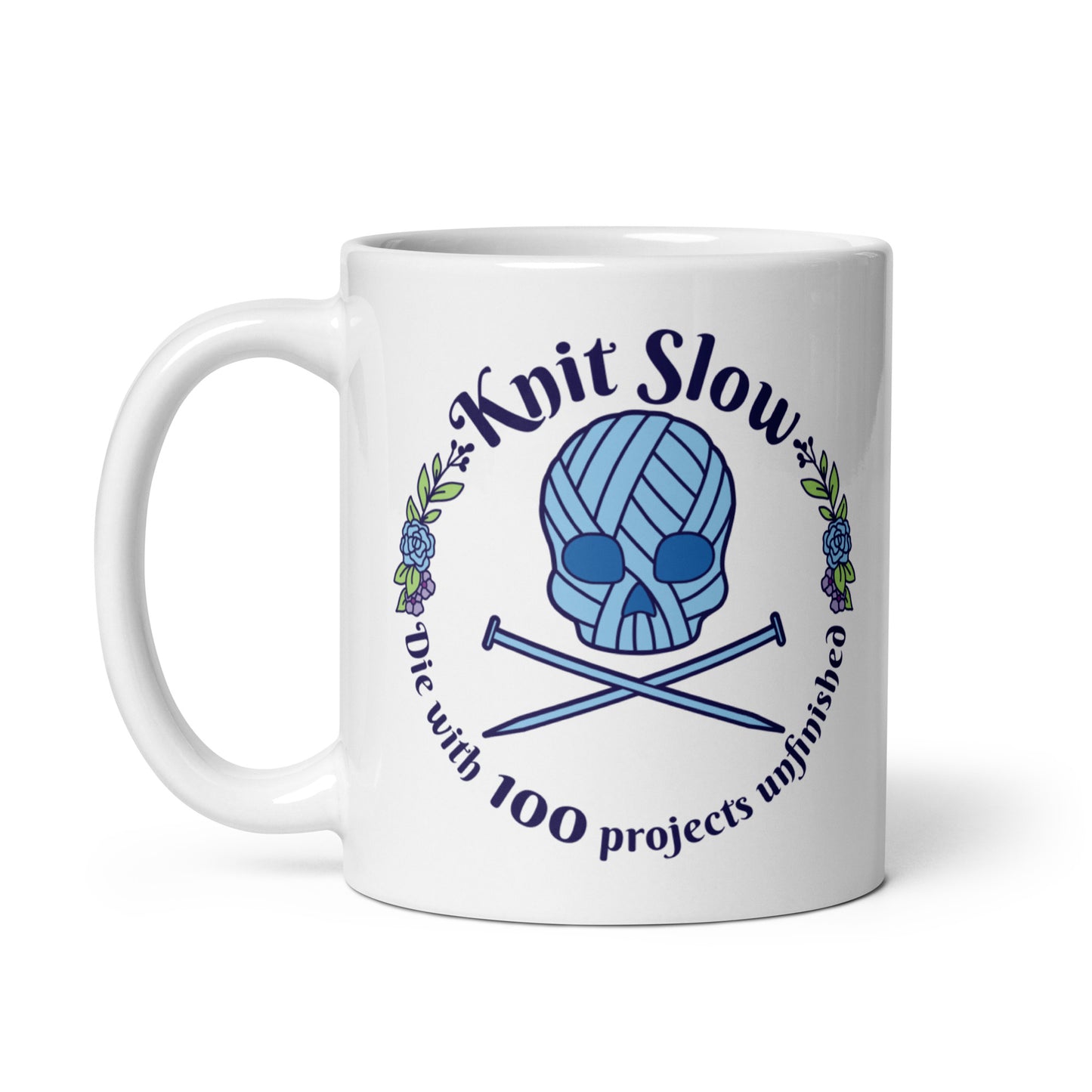 A white 11 ounce ceramic mug featuring an image of a skull and crossbones made of yarn and knitting needles. A floral wreath surrounds the skull, along with words that read "Knit Slow, Die with 100 projects unfinished"