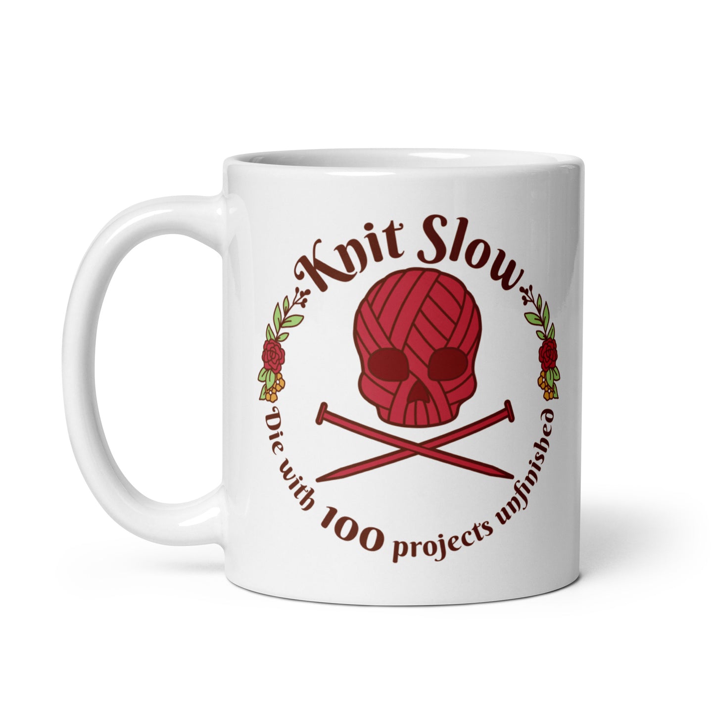 A white 11 ounce ceramic mug featuring an image of a skull and crossbones made of yarn and knitting needles. A floral wreath surrounds the skull, along with words that read "Knit Slow, Die with 100 projects unfinished"