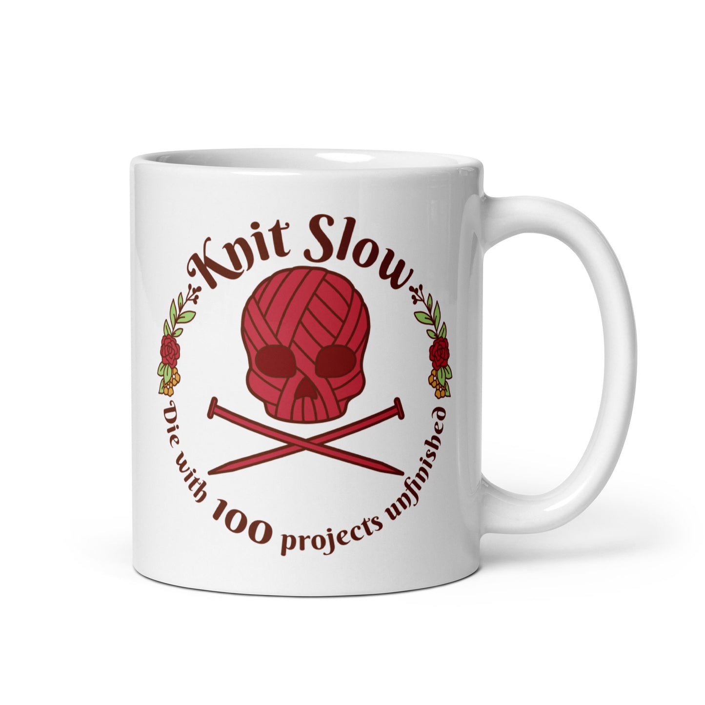 A white 11 ounce ceramic mug featuring an image of a skull and crossbones made of yarn and knitting needles. A floral wreath surrounds the skull, along with words that read "Knit Slow, Die with 100 projects unfinished"