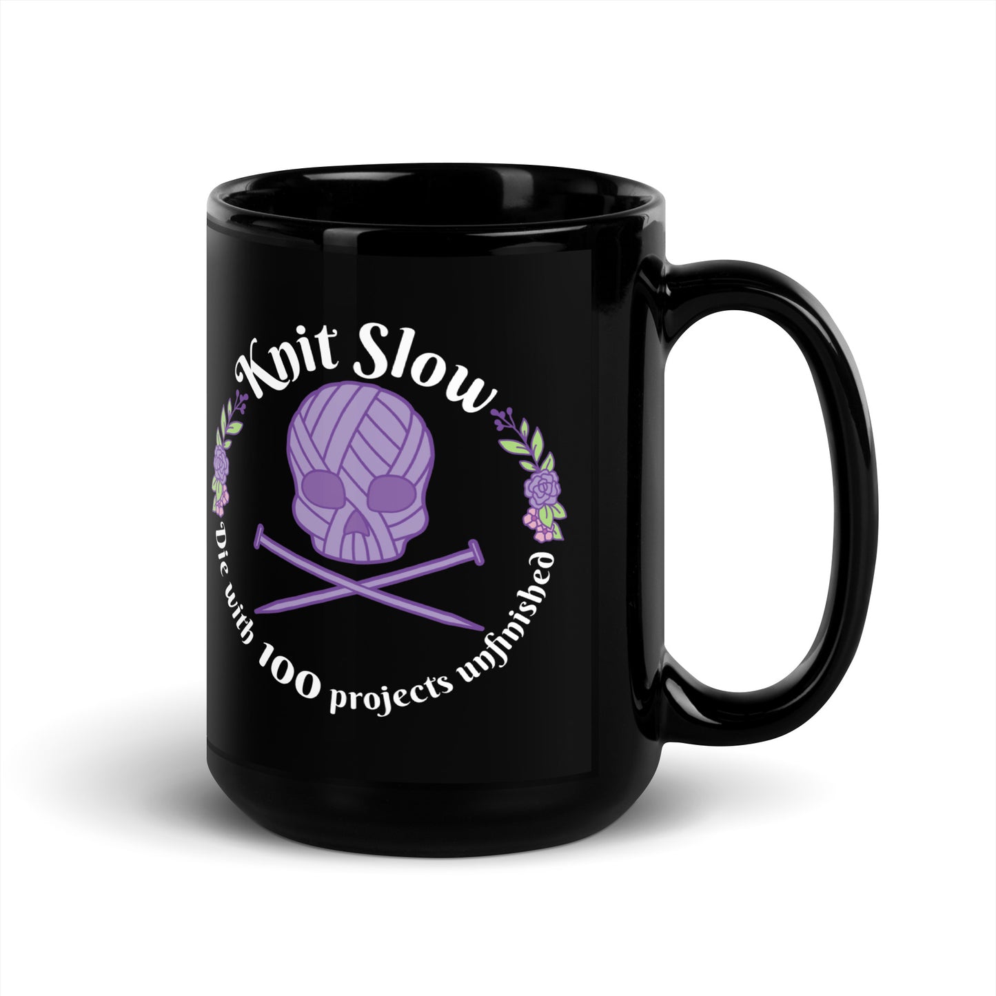 A black 15 ounce ceramic mug featuring an image of a skull and crossbones made of yarn and knitting needles. A floral wreath surrounds the skull, along with words that read "Knit Slow, Die with 100 projects unfinished"