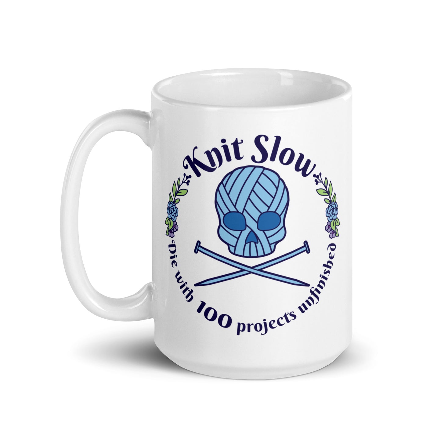 A white 15 ounce ceramic mug featuring an image of a skull and crossbones made of yarn and knitting needles. A floral wreath surrounds the skull, along with words that read "Knit Slow, Die with 100 projects unfinished"