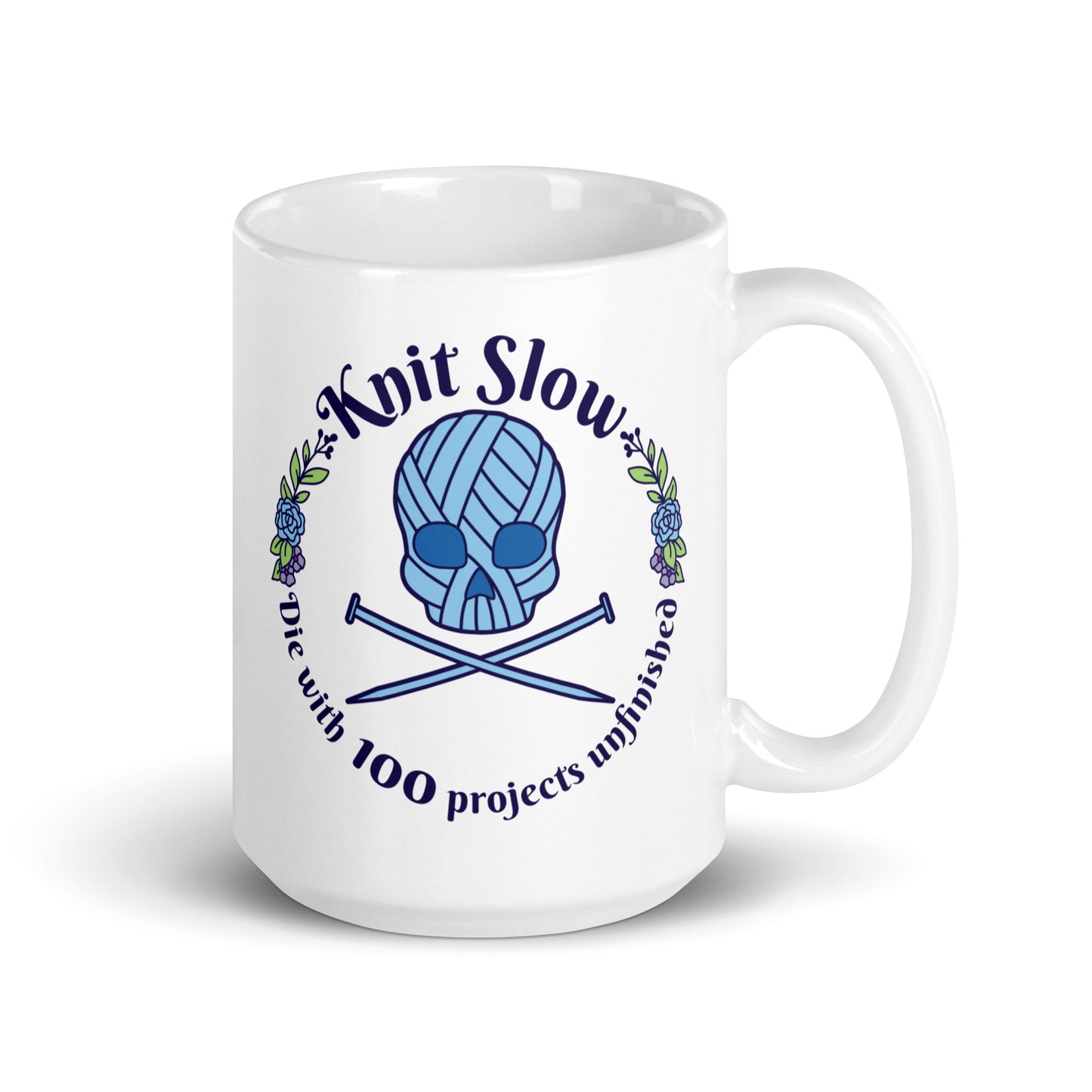 A white 15 ounce ceramic mug featuring an image of a skull and crossbones made of yarn and knitting needles. A floral wreath surrounds the skull, along with words that read "Knit Slow, Die with 100 projects unfinished"