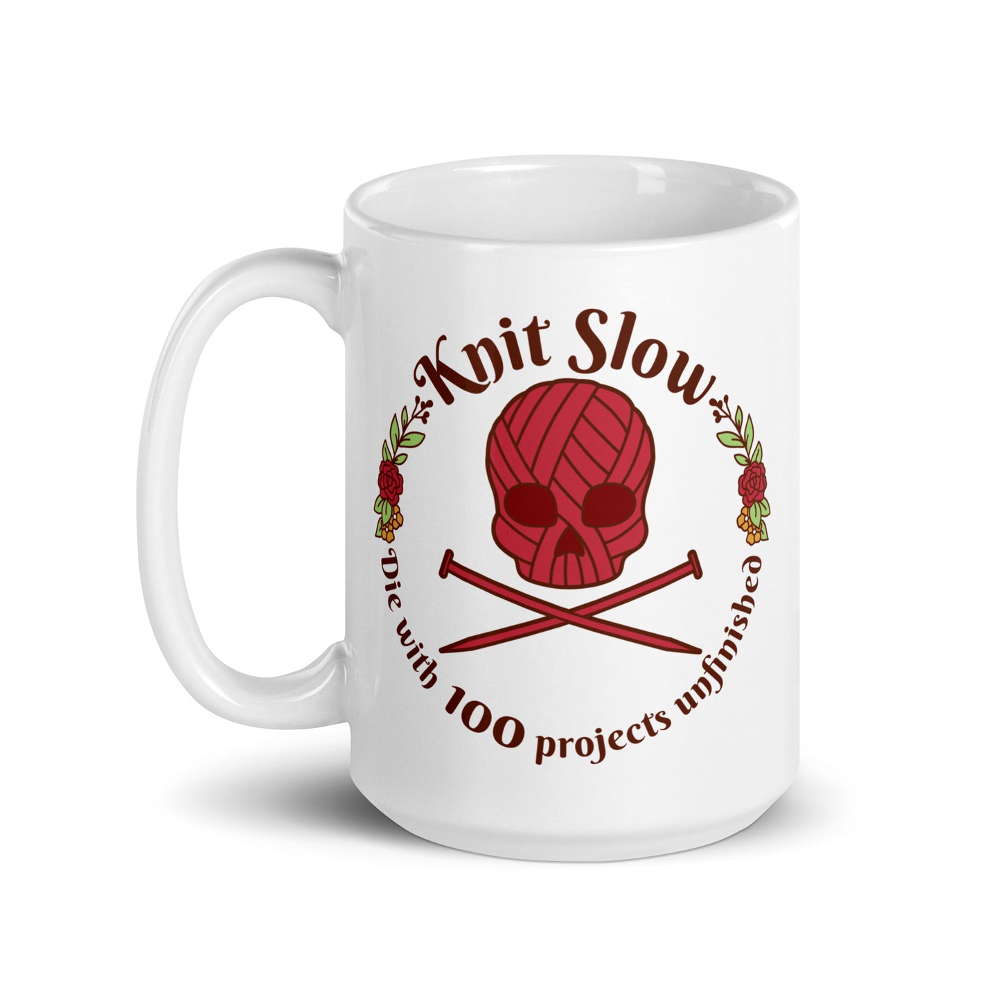 A white 15 ounce ceramic mug featuring an image of a skull and crossbones made of yarn and knitting needles. A floral wreath surrounds the skull, along with words that read "Knit Slow, Die with 100 projects unfinished"