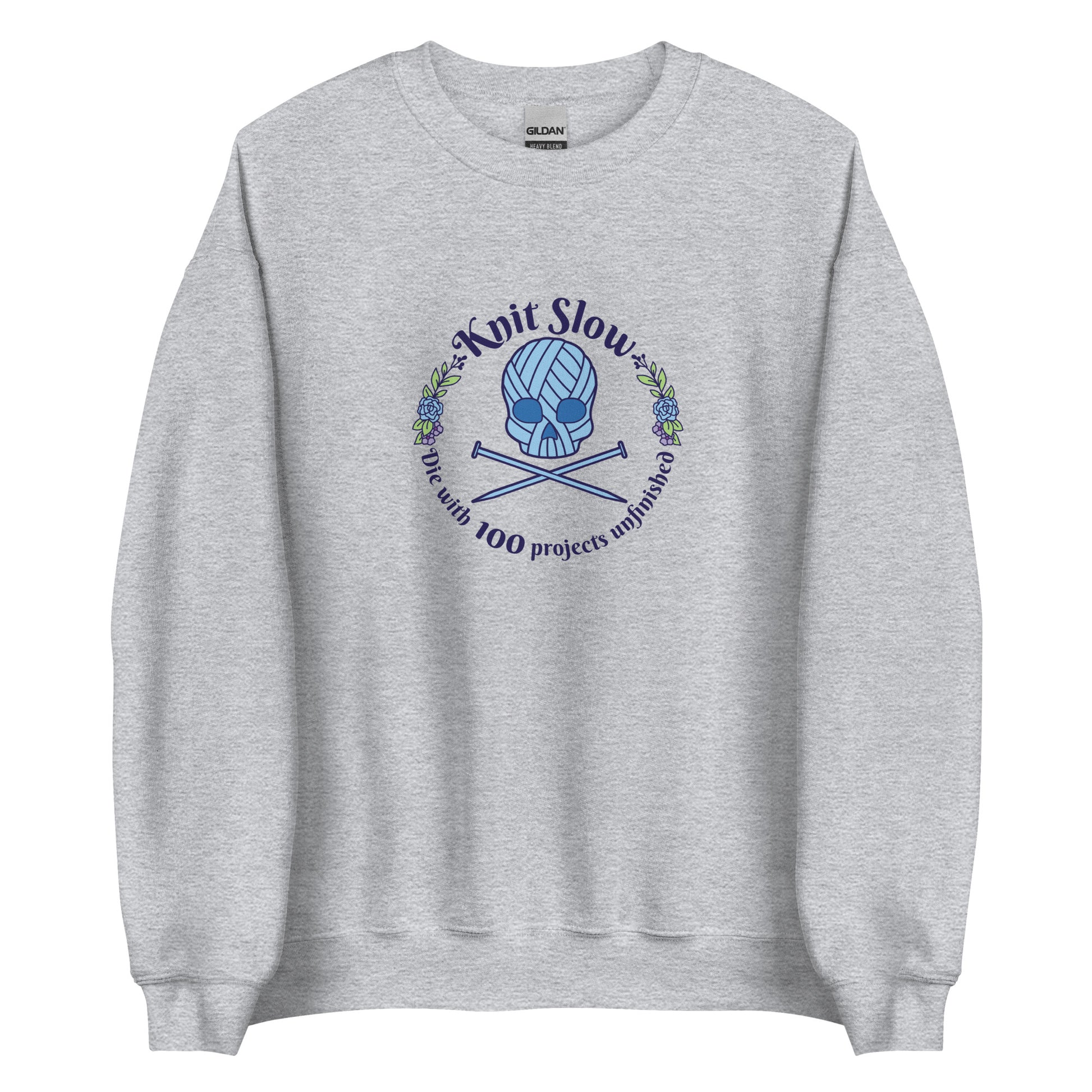 A grey crewneck sweatshirt featuring an image of a skull and crossbones made of yarn and knitting needles. A floral wreath surrounds the skull, along with words that read "Knit Slow, Die with 100 projects unfinished"