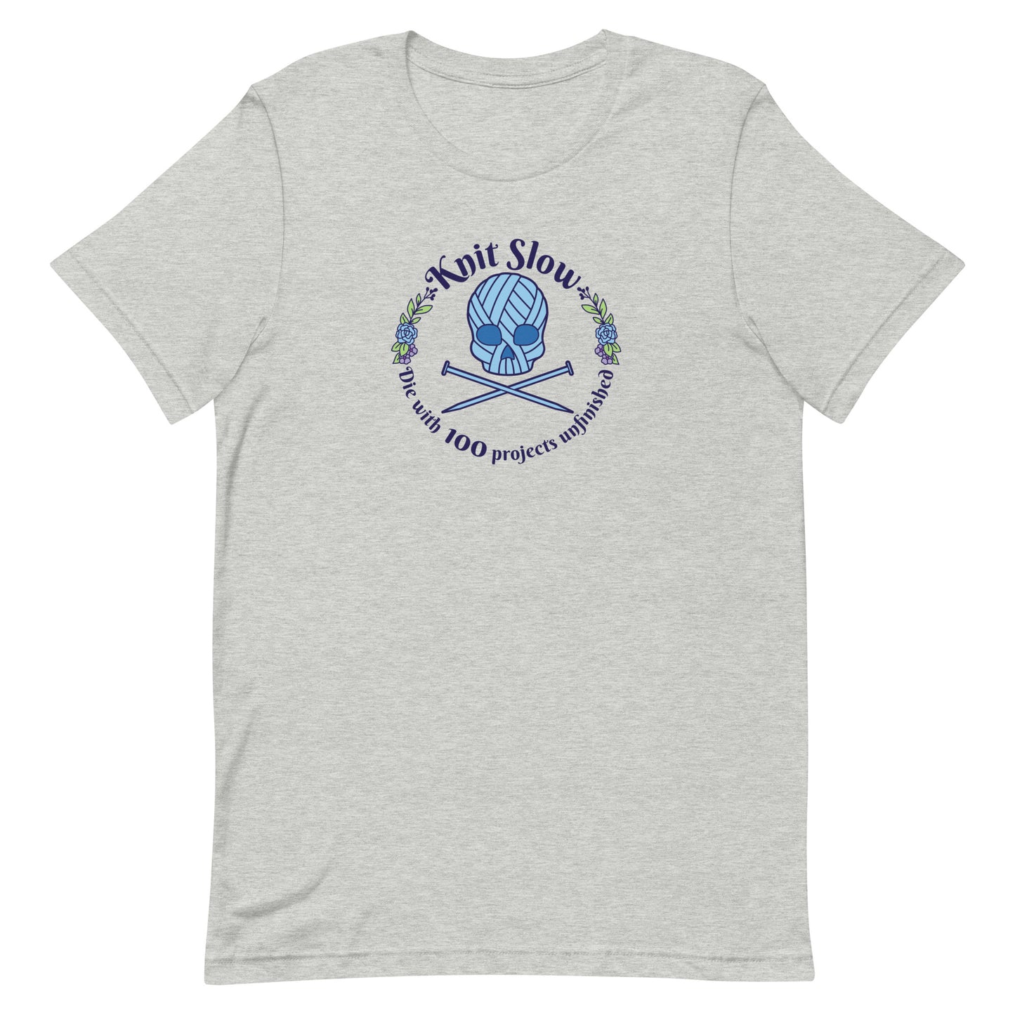 A heathered grey crewneck t-shirt featuring an image of a skull and crossbones made of yarn and knitting needles. A floral wreath surrounds the skull, along with words that read "Knit Slow, Die with 100 projects unfinished"