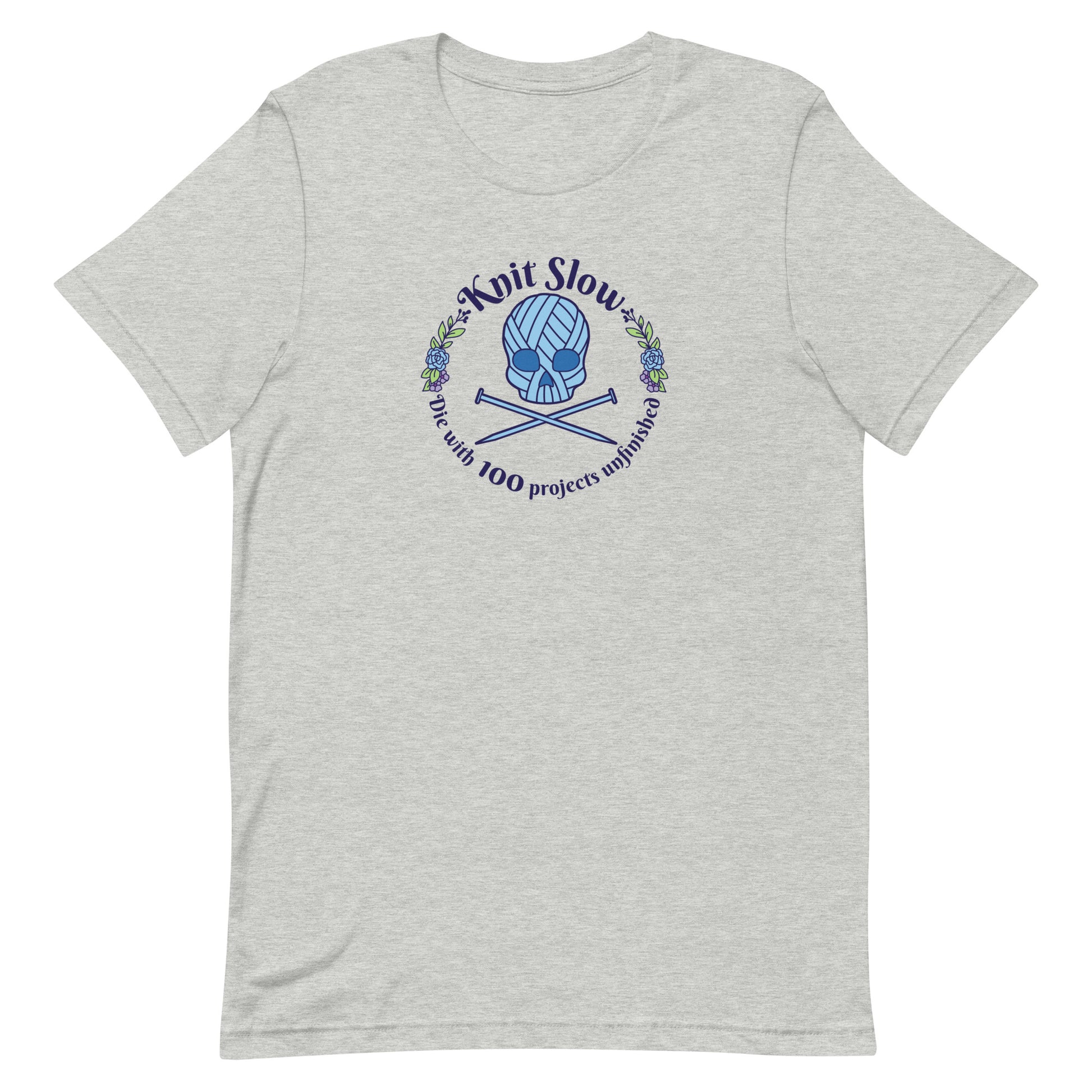 A heathered grey crewneck t-shirt featuring an image of a skull and crossbones made of yarn and knitting needles. A floral wreath surrounds the skull, along with words that read "Knit Slow, Die with 100 projects unfinished"