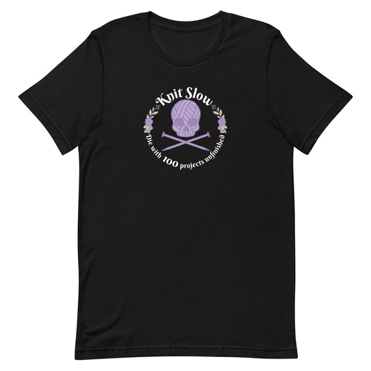 A black crewneck t-shirt featuring an image of a skull and crossbones made of yarn and knitting needles. A floral wreath surrounds the skull, along with words that read "Knit Slow, Die with 100 projects unfinished"