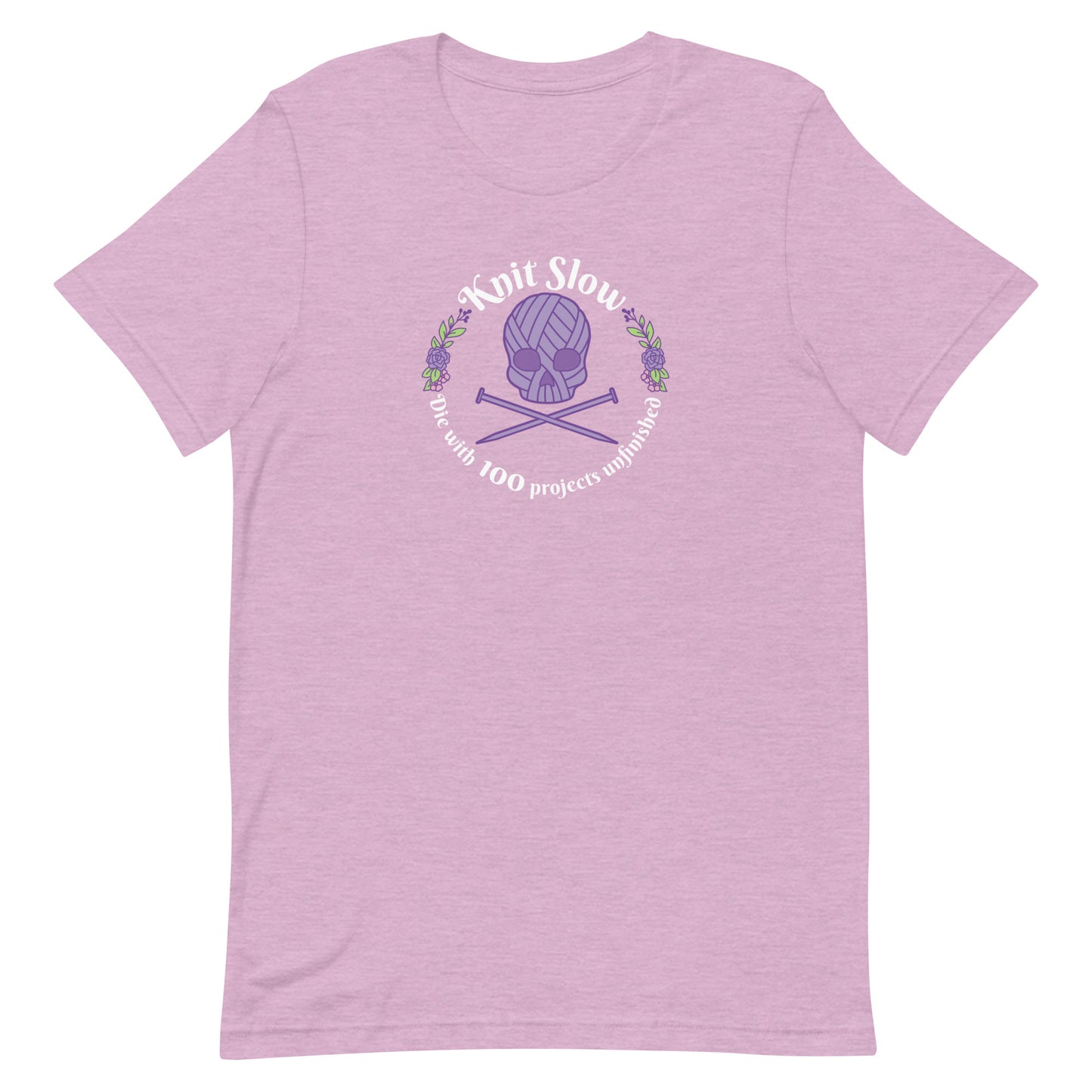 A heathered pink crewneck t-shirt featuring an image of a skull and crossbones made of yarn and knitting needles. A floral wreath surrounds the skull, along with words that read "Knit Slow, Die with 100 projects unfinished"