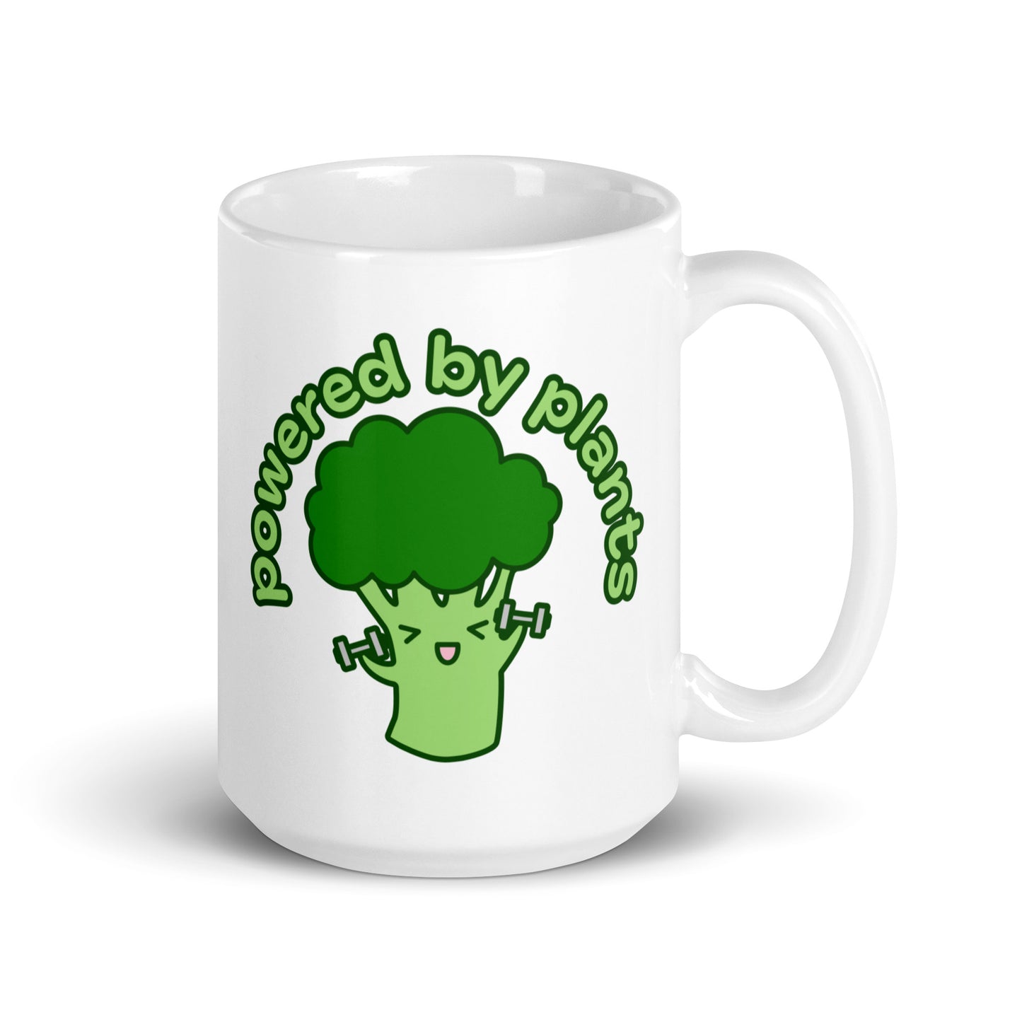 A white 15 ounce ceramic coffee mug featuring a cute illustration of a cartoon broccoli. The broccoli is excitedly lifting weights and text above him in an arc reads "powered by plants".
