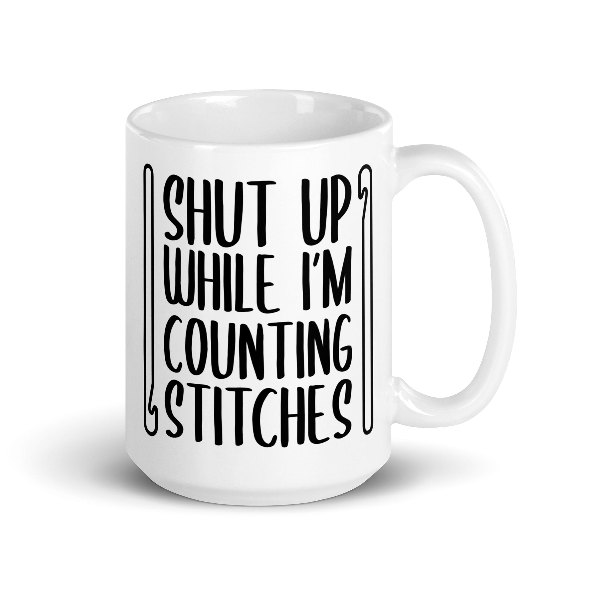 A white 15 ounce ceramic mug featuring black text that reads "Shut up while I'm counting stitches." The text is framed by a crochet hook to the left and right.