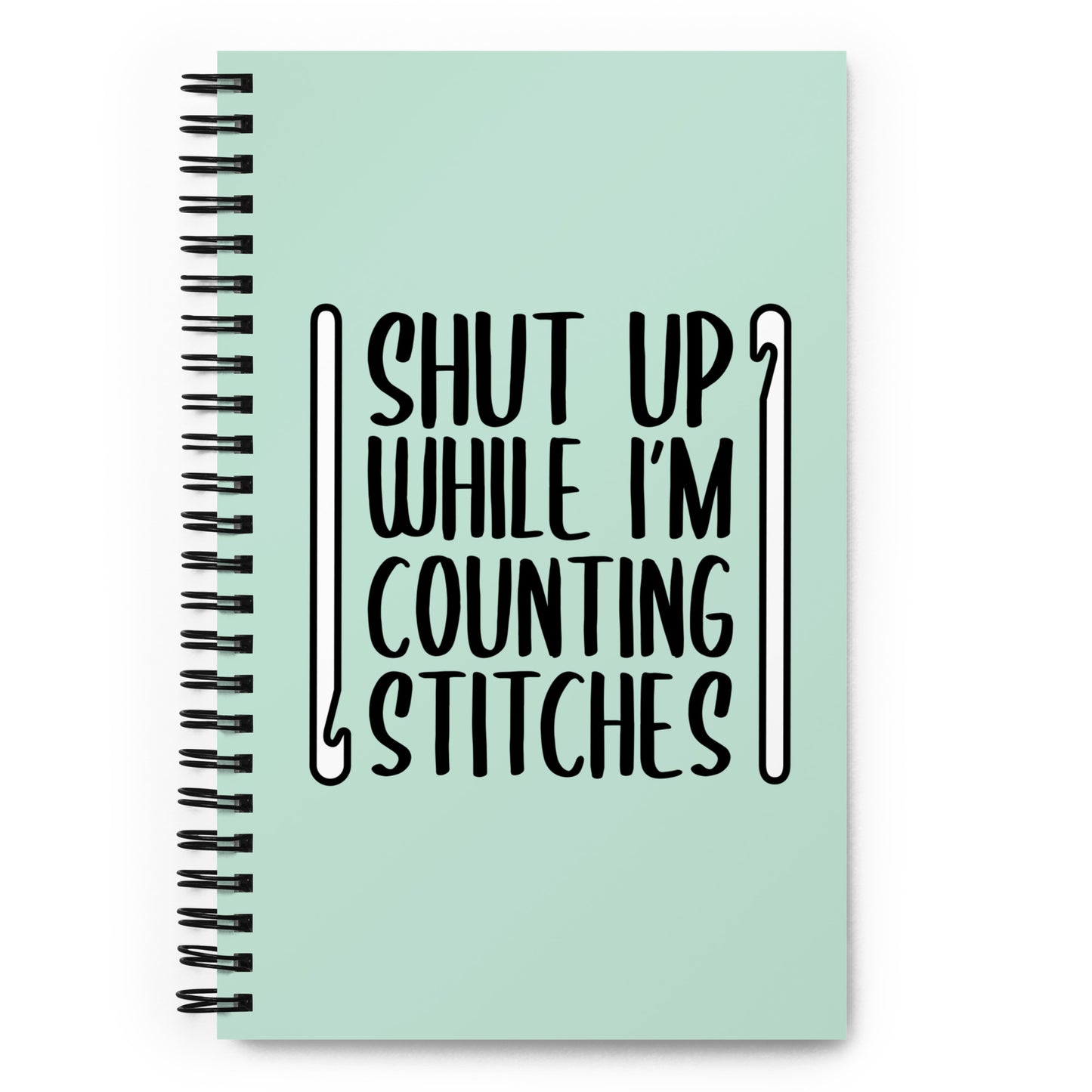 A mint green o-ring bound notebook featuring black text that reads "Shut up while I'm counting stitches." The text is framed by a crochet hook to the left and right.