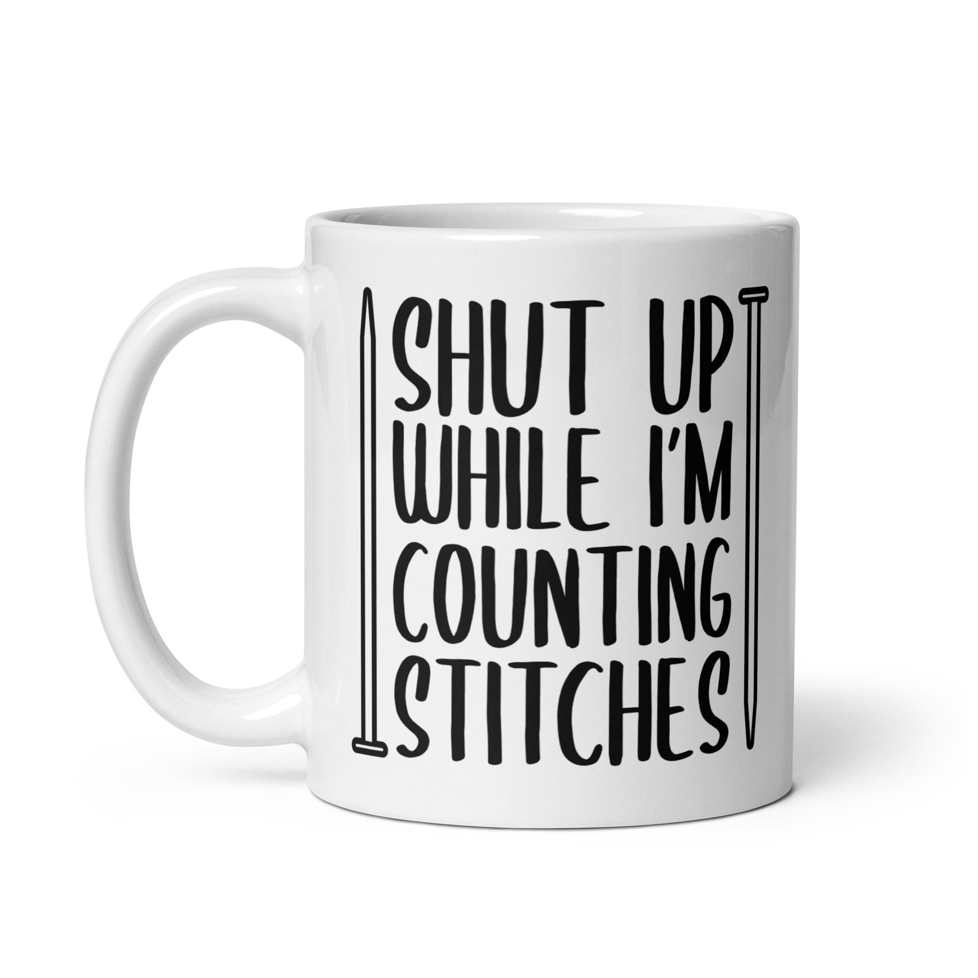 A white 11 ounce ceramic mug with black text surrounded by two knitting needles. The text reads "Shut up while I'm counting stiches".