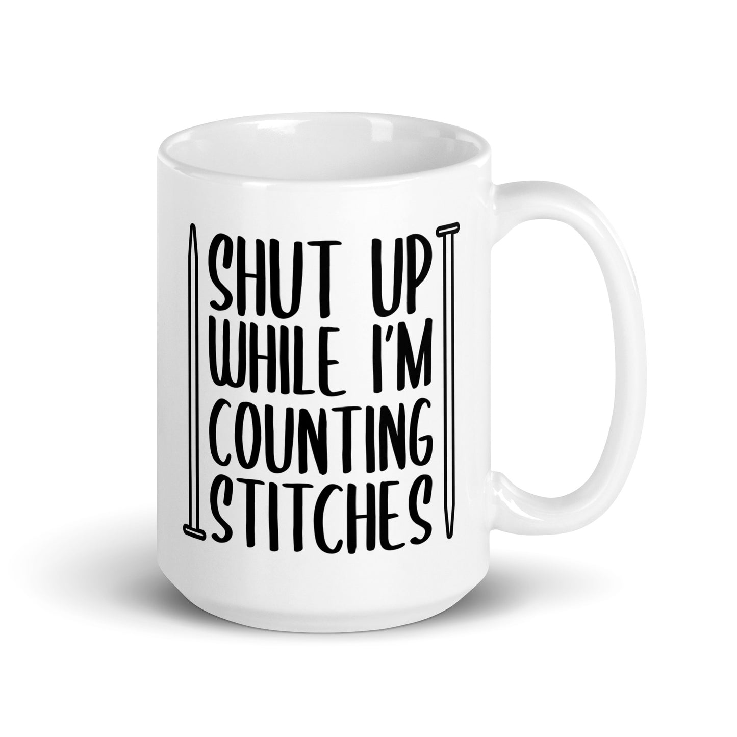 A white 15 ounce ceramic mug with black text surrounded by two knitting needles. The text reads "Shut up while I'm counting stiches".