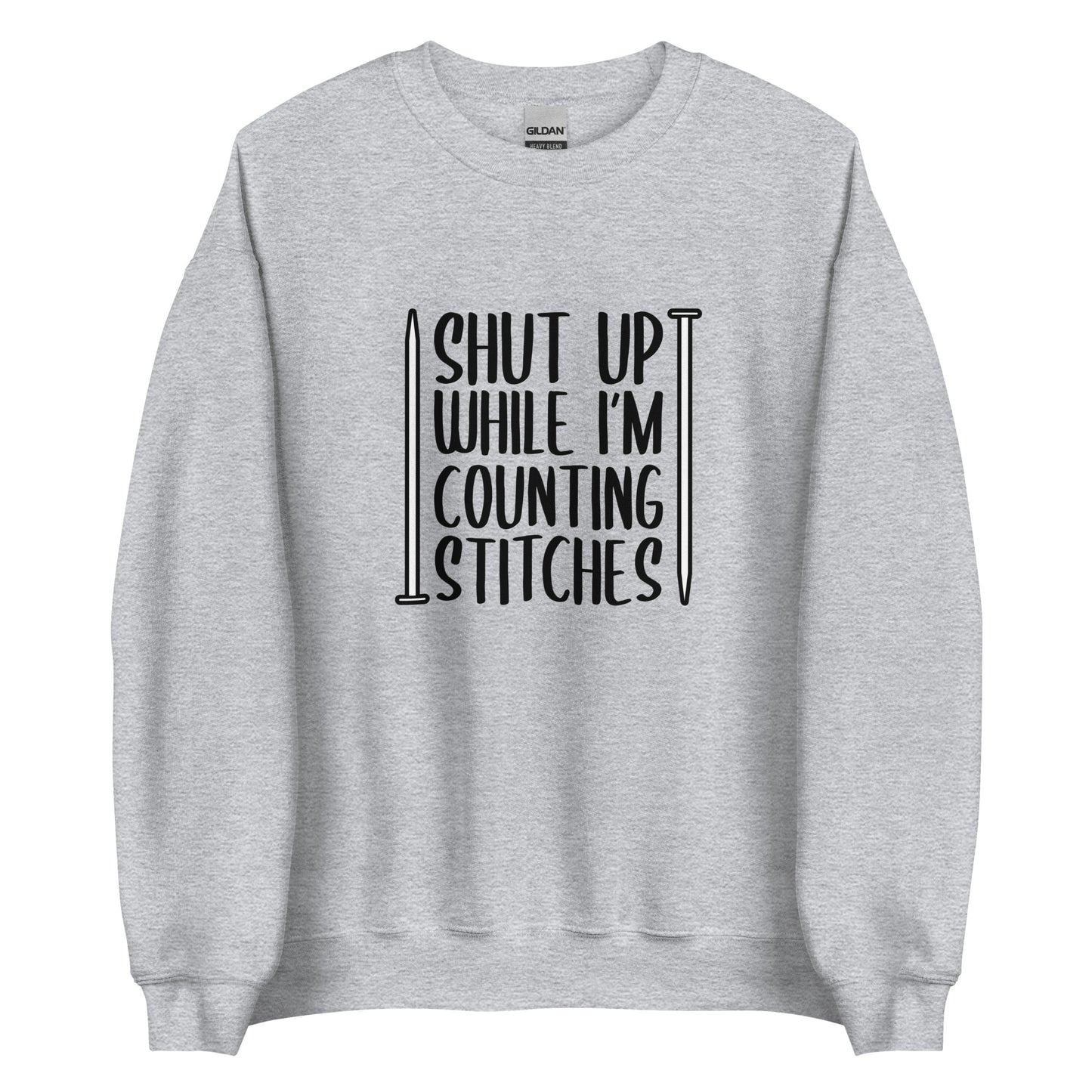 A grey crewneck sweatshirt with black text surrounded by two knitting needles. The text reads "Shut up while I'm counting stiches".
