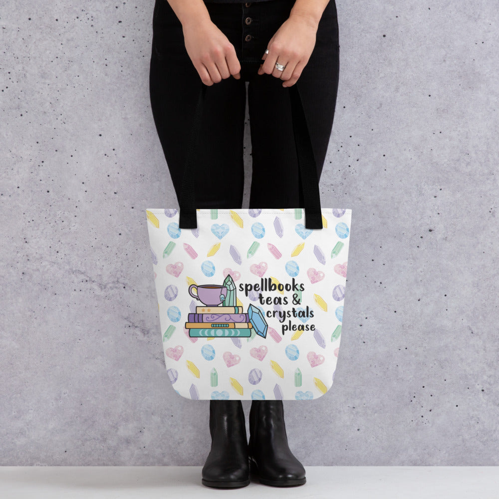 A waist-down image of a person holding a white totebag with black handles, decorated with a pattern of pastel-colored crystals. An illustration on the bag shows a stack of spellbooks with a teacup on top and crystals scattered around the books. Text next to the image reads "Spellbooks, teas & crystals please"