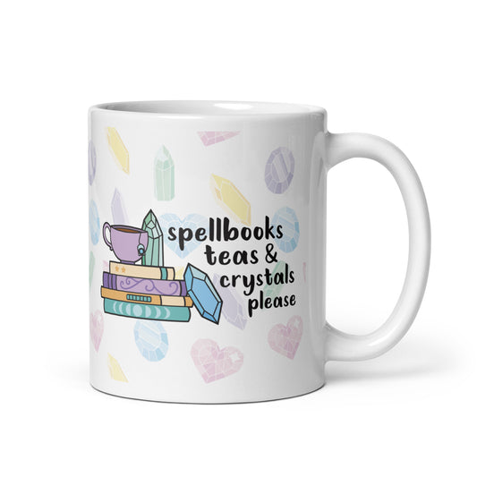 A white 11 ounce ceramic mug with a pattern of pastel crystals. In the center of the mug is an illustration of several books and crystals and a teacup. Text along side the image reads "Spellbooks, teas, & crystals please"