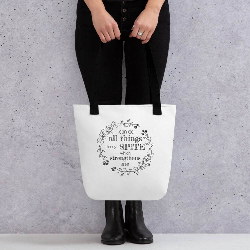 A waist-down image of a model wearing all black who is holding a white tote bag with black handles, featuring a simple illustration of a floral wreath. Inside the wreath is text that reads "I can do all things through SPITE which motivates me"