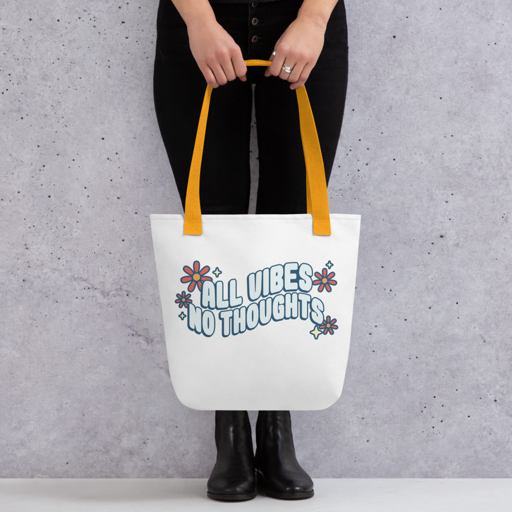 A waist-down image of a model wearing all black. The model is holding a white tote bag with yellow handles. On the tote bag is text that reads "al vibes no thoughts" with flowers and sparkles.