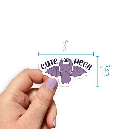 A hand holding a diecut sticker featuring a cutesy, smiling purple bat. Text above the bat's wings reads "Cute as heck". Measurements around the sticker read "3 inches wide" by "1.6 inches tall"