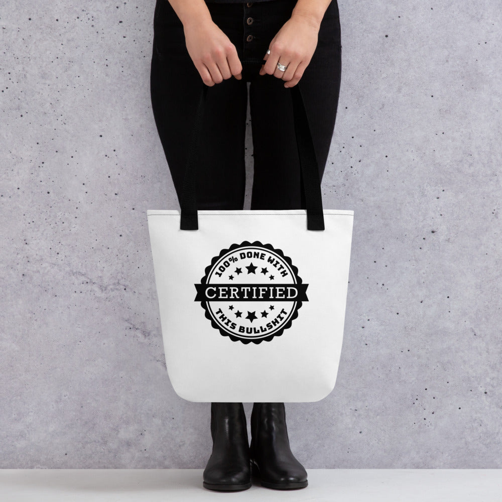 A waist-down image of a model who is wearing all black. The model is holding a white tote bag with black handles and stitching. Centered on the bag is an image of an official-looking stamped seal that reads "CERTIFIED: 100% done with this bullshit."