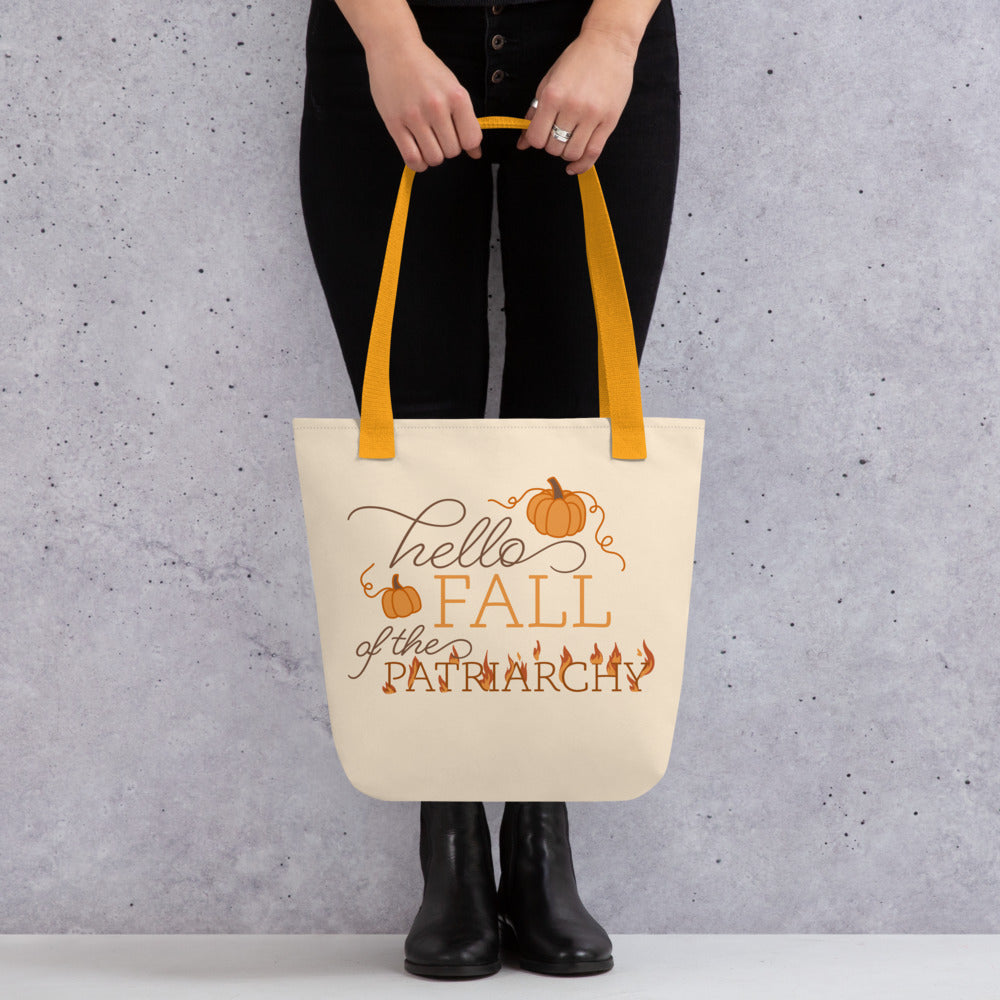 A waist-down image of a model wearing all black, and holding a tote bag. The tote bag is cream-colored, and features text in a blend of cursive and serif fonts that read "hello FALL of the PATRIARCHY". Pumpkins surround the upper half of the text, and the word "Patriarchy" is surrounded by flames.