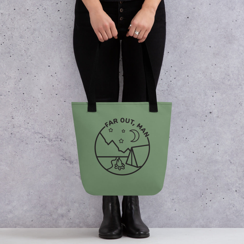 A waist-down image of a model wearing all black holding an olive green tote bag with black handles. The tote bag is decorated with a black lineart illustration of a campfire and tent under a night sky. Text in an arc above the illustration reads "Far out, man".