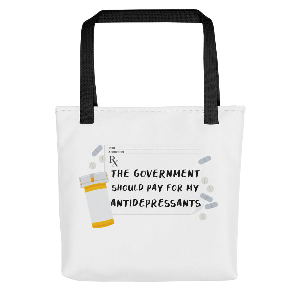A white tote bag with black handles. On the tote bag is an image of a prescription sheet surrounded by an orange pill bottle and pills of various colors and shapes. Text on the prescription reads "The government should pay for my antidepressants"