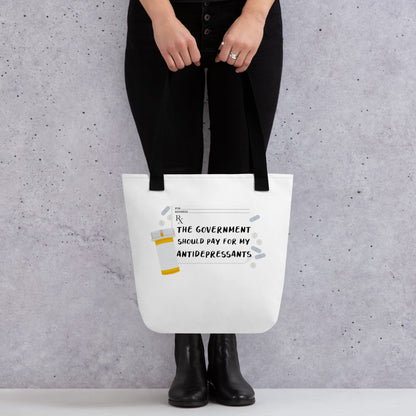 A waist-down image of a model wearing all black, who is holding A white tote bag with black handles. On the tote bag is an image of a prescription sheet surrounded by an orange pill bottle and pills of various colors and shapes. Text on the prescription reads "The government should pay for my antidepressants"