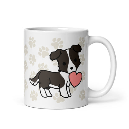 A white 11 ounce ceramic mug with a pattern of light brown pawprints all over. The mug also features a stylized illustration of a Border Collie holding a heart in its mouth.