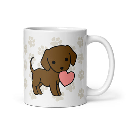A white 11 ounce ceramic mug with a pattern of light brown pawprints all over. The mug also features a stylized illustration of a Chocolate Lab holding a heart in its mouth.