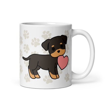 A white 11 ounce ceramic mug with a pattern of light brown pawprints all over. The mug also features a stylized illustration of a Rottweiler holding a heart in its mouth.