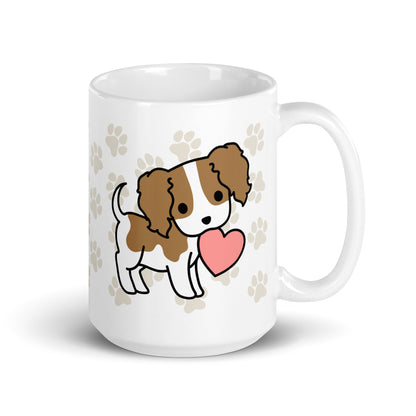 A white 15 ounce ceramic mug with a pattern of light brown pawprints all over. The mug also features a stylized illustration of a Cavalier King Charles Spaniel holding a heart in its mouth.