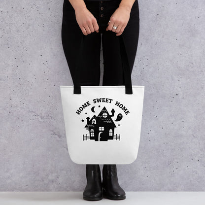A waist-down image of a model who is wearing all black. The model is holding a white tote bag with black stitching and handles. Featured on the bag is a black silhouette of a haunted house. Text above the house reads "Home Sweet Home"